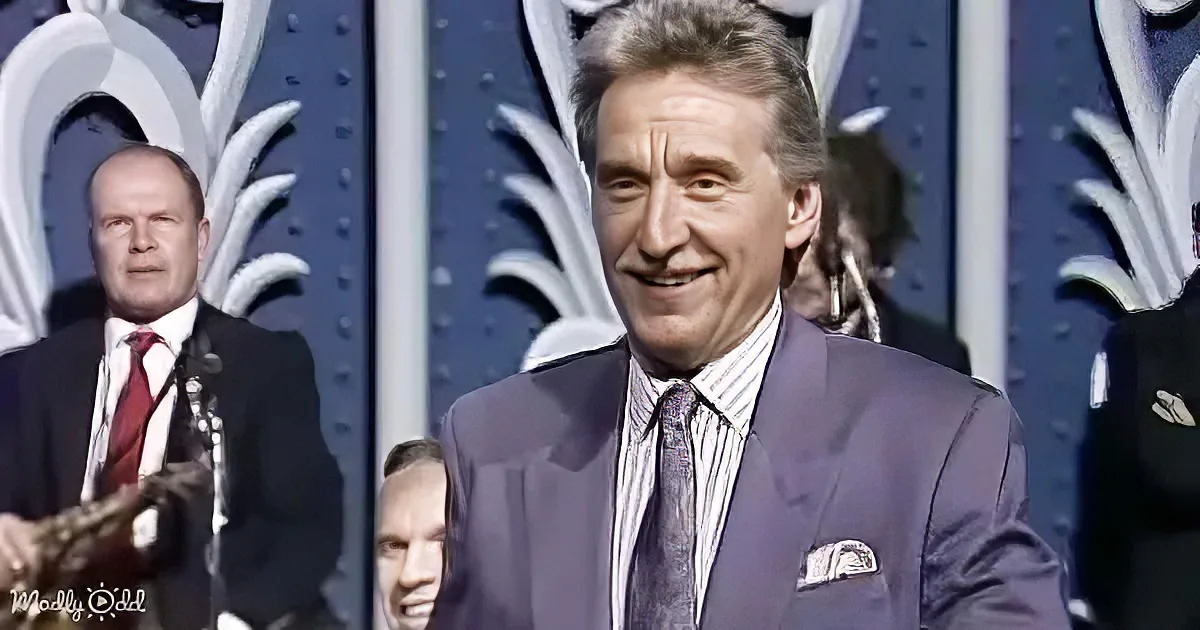 A man in a suit and tie smiles on stage, with another man in a suit and red tie behind him.