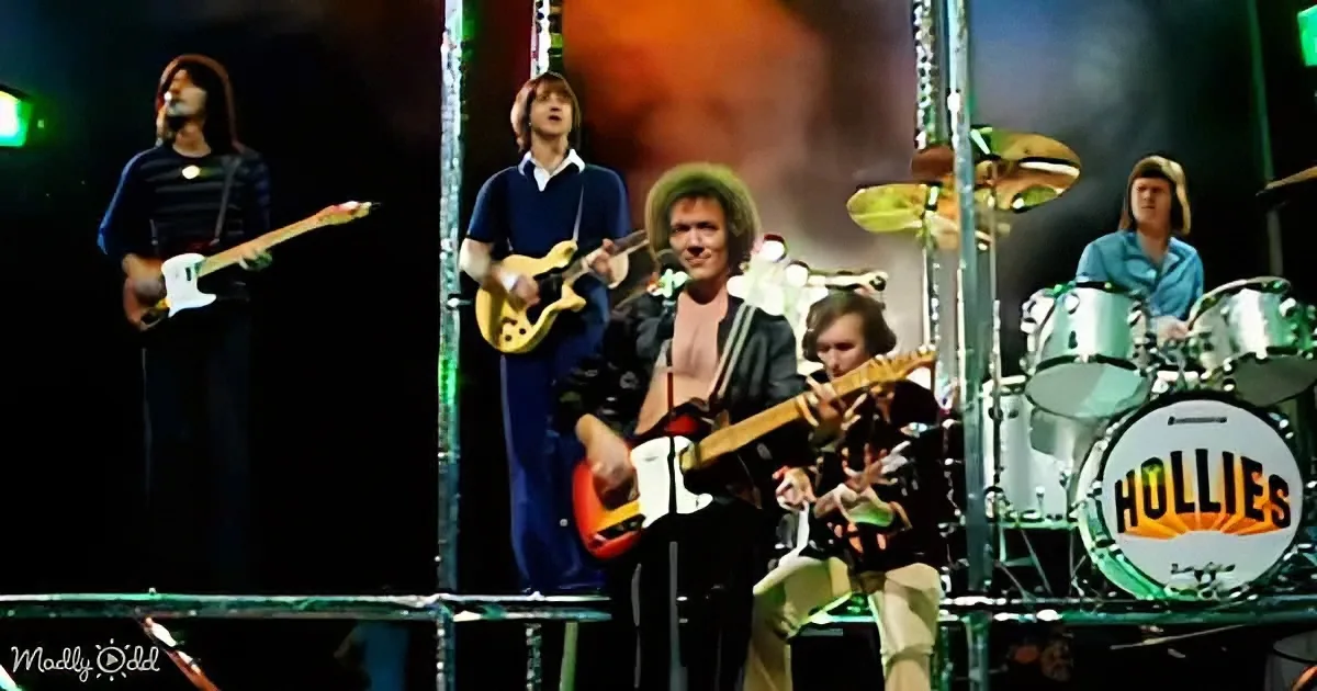 The Hollies band performs on stage with guitars, drums, and a bassist, illuminated by stage lights.