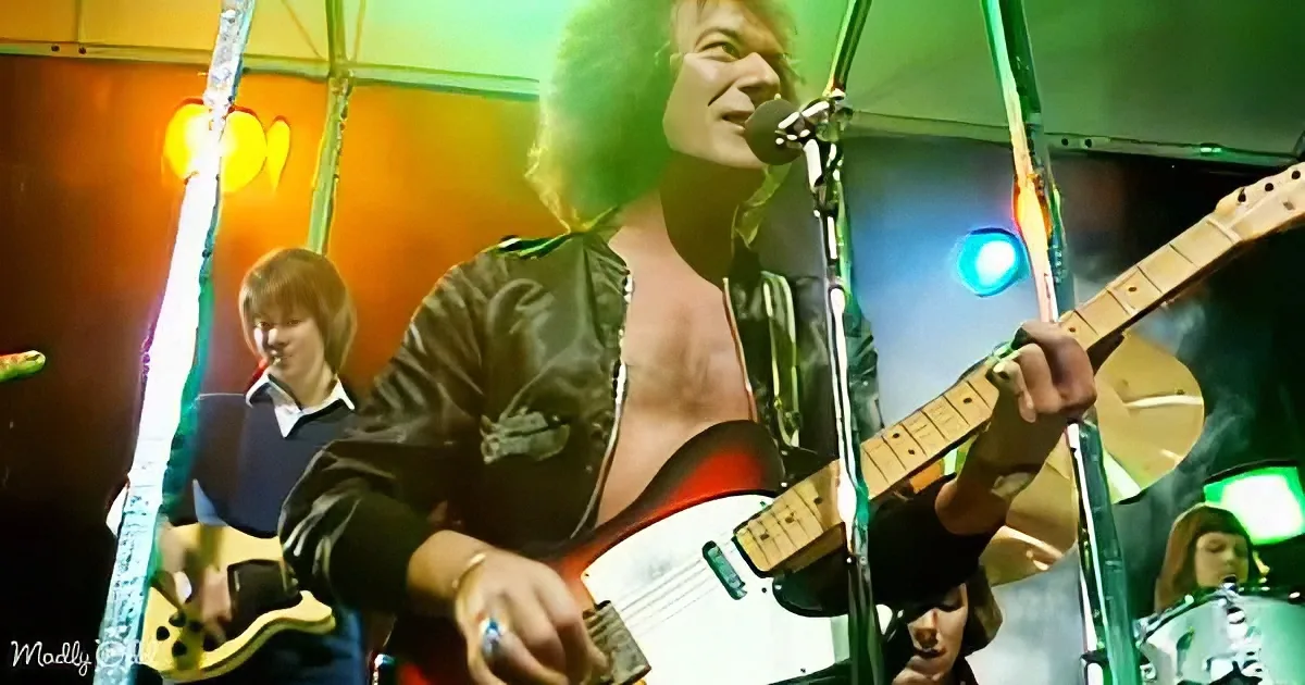 A man with curly hair plays guitar on stage, wearing a black jacket, with a microphone in front of him.