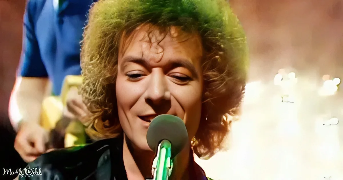 A person with curly green hair sings into a microphone, with a guitarist in the background.