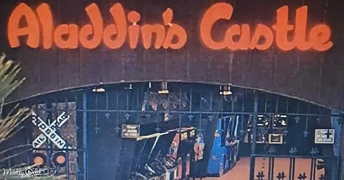 A sign reading "Aladdin's Castle" in orange letters atop a dark structure with a gate and a train crossing sign.