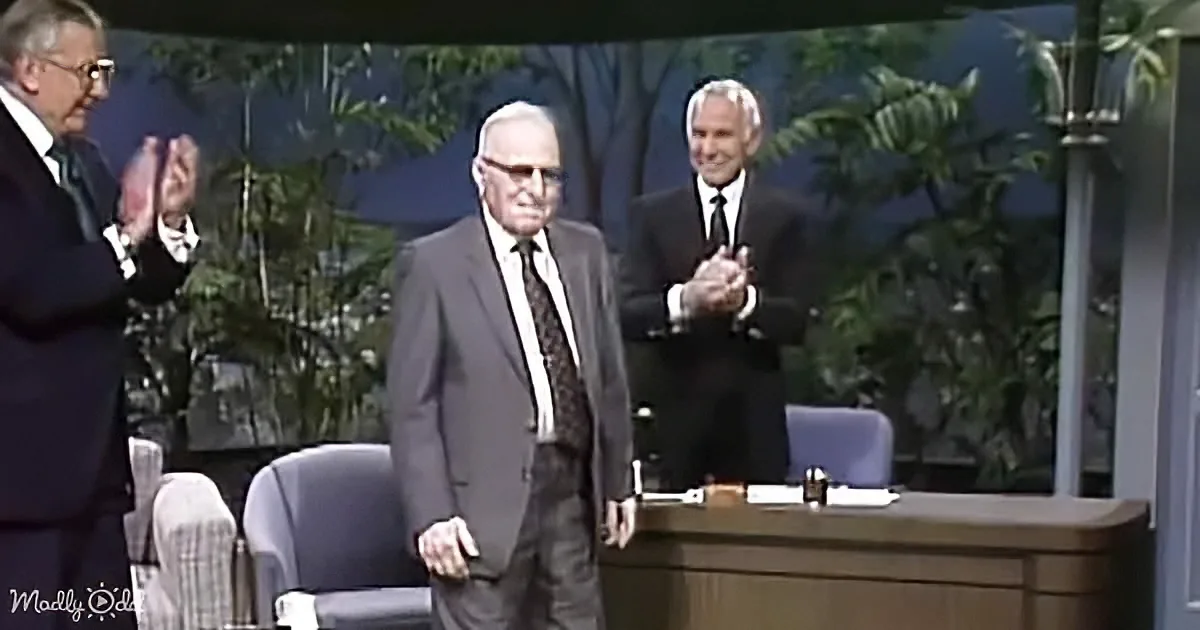 An older man in a suit stands on a stage, clapping with two other men in suits behind him.
