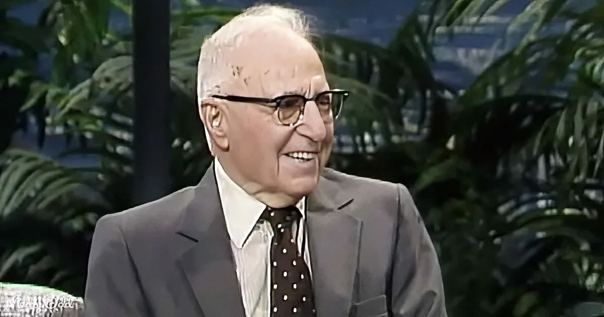 An older man in a suit and tie smiles while wearing glasses, with a tropical background.