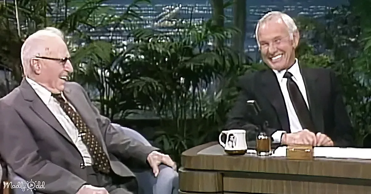Two men in suits, one seated, one standing, smiling, with a backdrop of greenery and a table with mugs and a box.