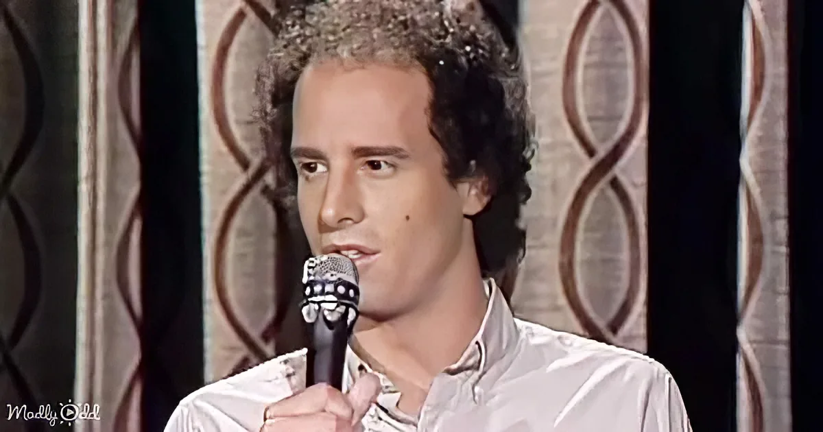 A man with curly hair, wearing a white shirt, holds a microphone and appears to be speaking or singing.
