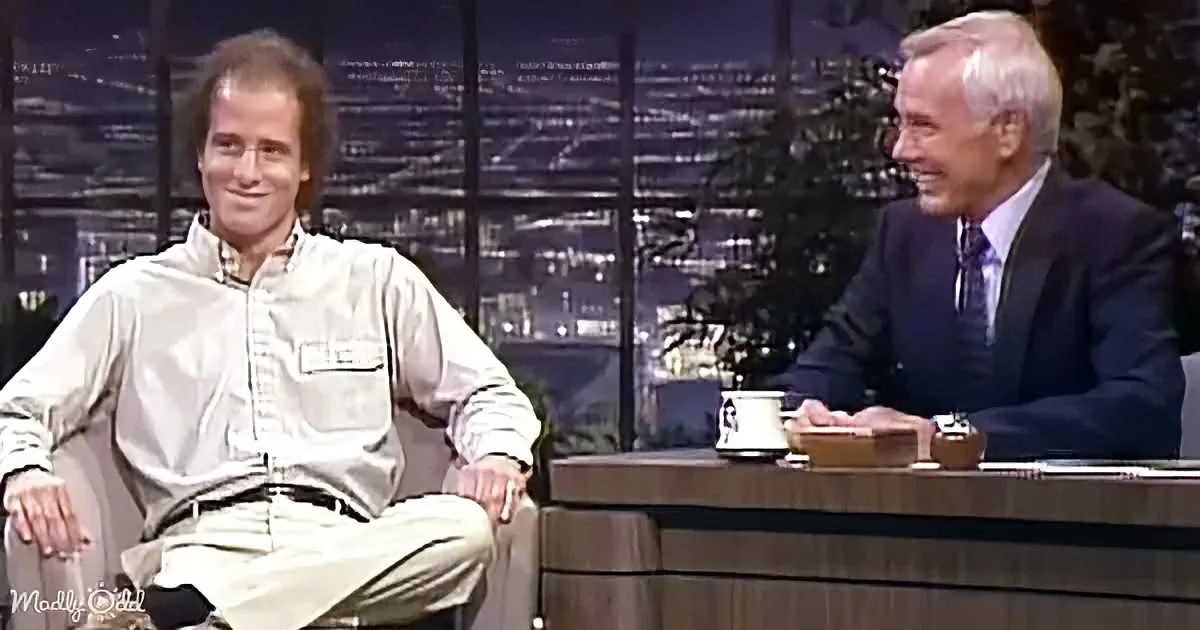 A man in a light shirt and khaki pants sits on a couch, smiling, while another man in a suit sits behind a desk, also...