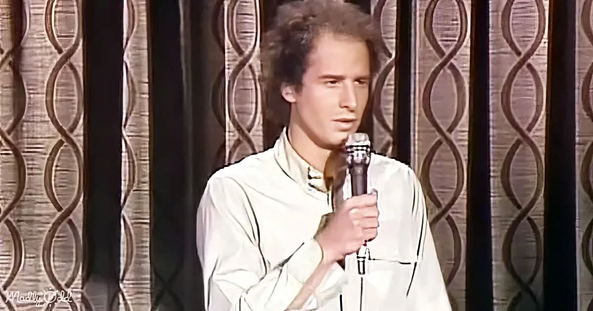 A man in a white shirt holds a microphone, standing in front of a patterned curtain.