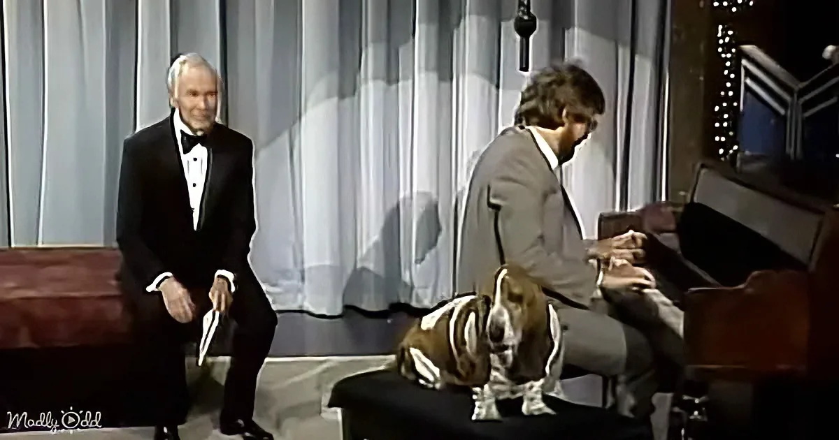 A man in a tuxedo stands beside a seated man at a piano, with a dog sitting between them on a stool.