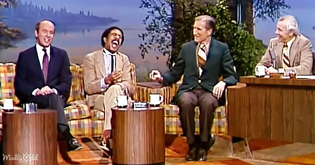 Four men in suits sit on a talk show set, laughing and smiling.