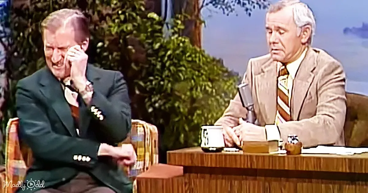 A man in a suit sits on a couch, covering his face, while another man in a suit sits at a desk with a mug and papers.