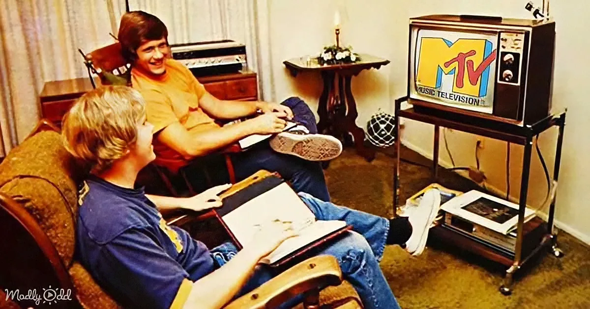 Two young men are sitting in a living room, one holding a book, watching an MTV-branded television.