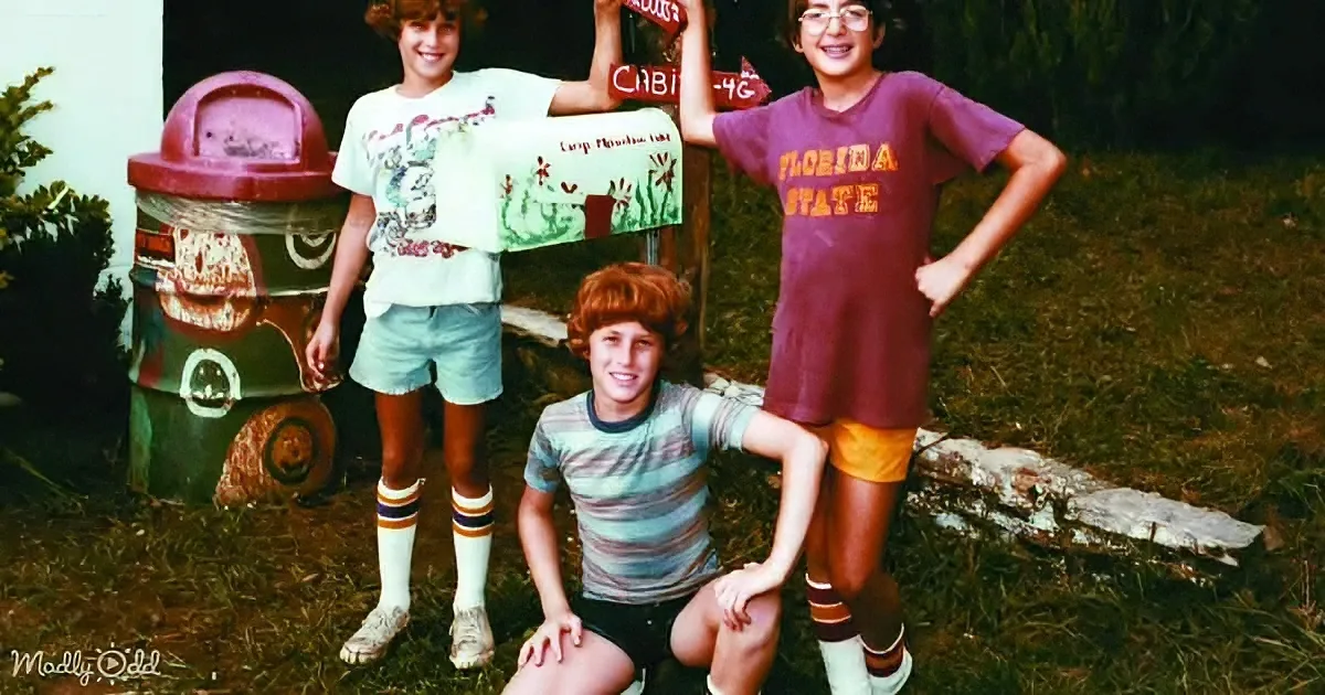 Three boys in retro clothing pose in front of a whimsical, painted trash can in a grassy yard.