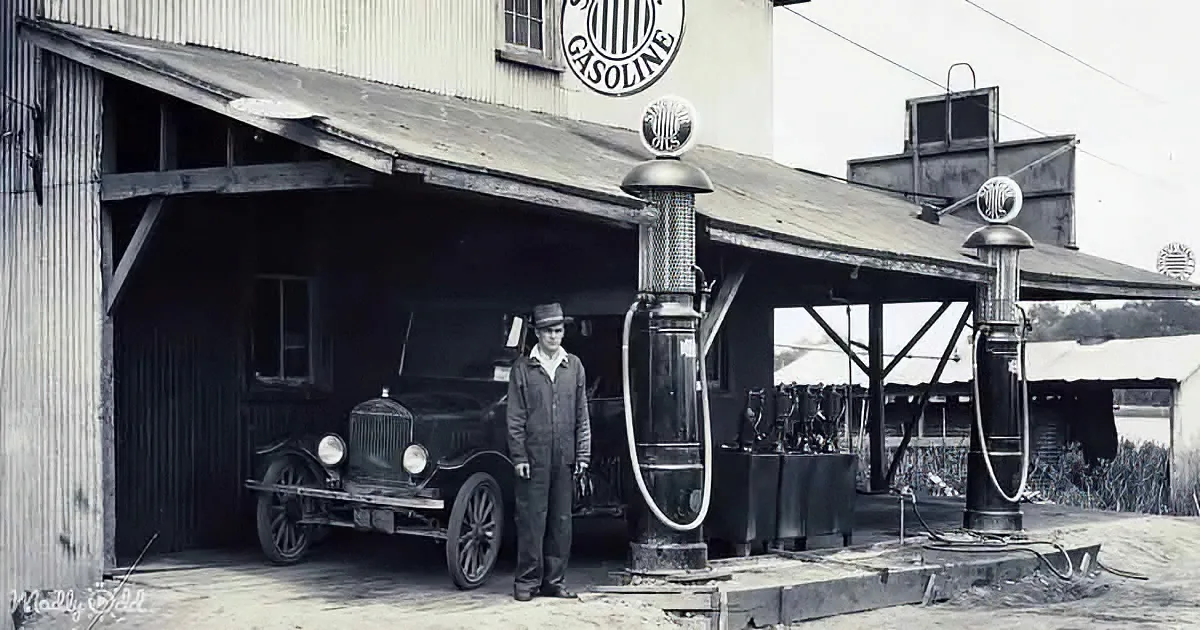 A man stands beside an old car at a gas station with vintage pumps and a sign reading "Gasoline."