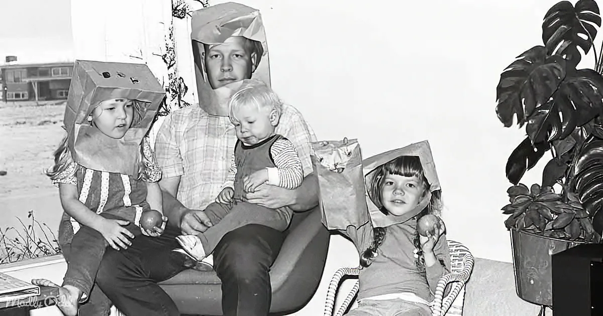 A man holds a baby while two children, wearing paper bag hats, sit on either side, holding apples.