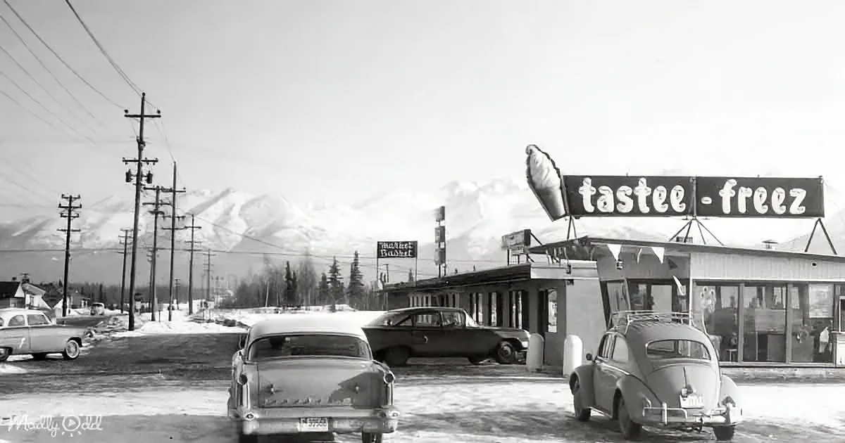 Vintage cars in front of a Tastee-Free ice cream shop with snow-covered mountains in the background.