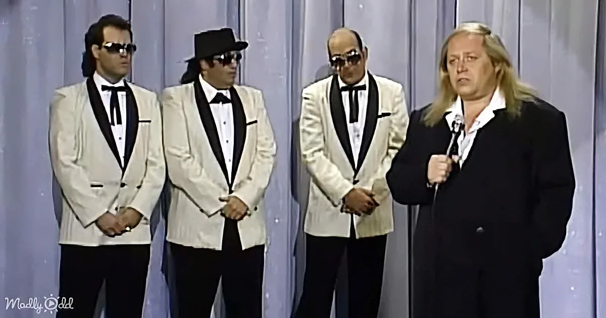 A man in a black suit holds a microphone, standing in front of three men in white suits with black lapels and sunglasses.