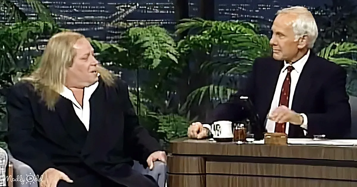 A man in a suit sits at a desk, gesturing, while another man in a suit leans forward, appearing to speak animatedly.
