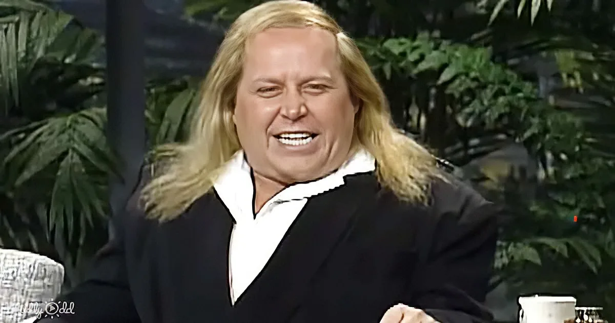 A person with long blonde hair, wearing a black suit and white shirt, is sitting with a hand on their hip, smiling.