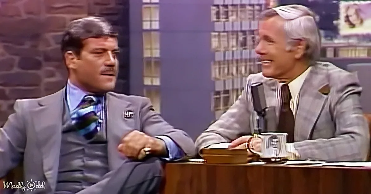 Two men in suits sit at a desk, one holding a mug, in a studio setting.