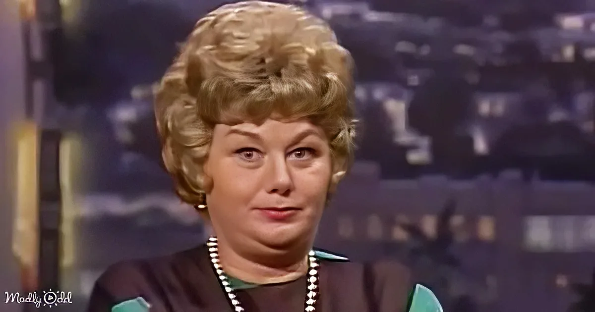 A woman with blonde, curly hair and a pearl necklace is seen against a blurred background.