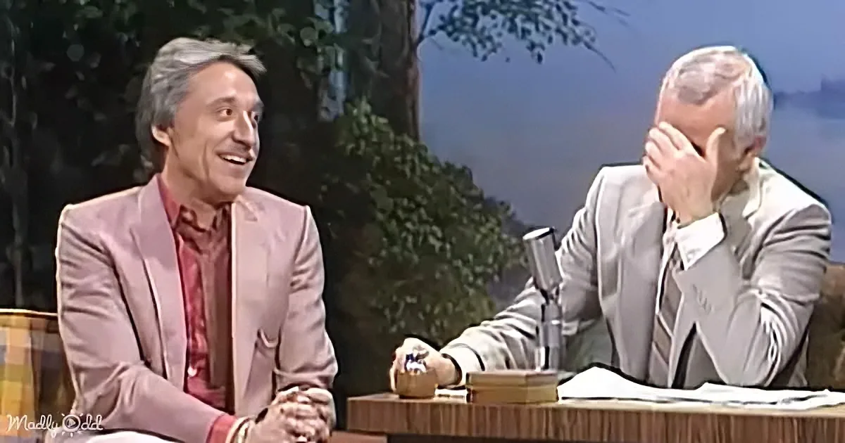 A man in a pink suit smiles while another in a white suit holds his head, both seated at a table.