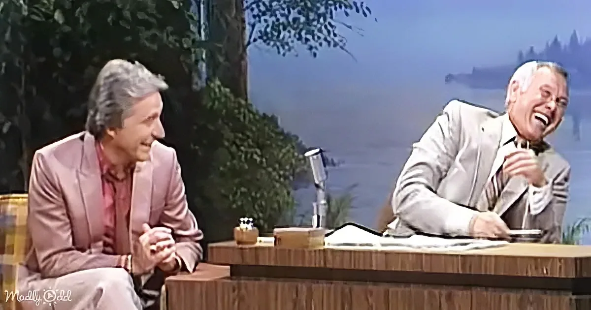 Two men are seated on a talk show set, one laughing while the other listens attentively.