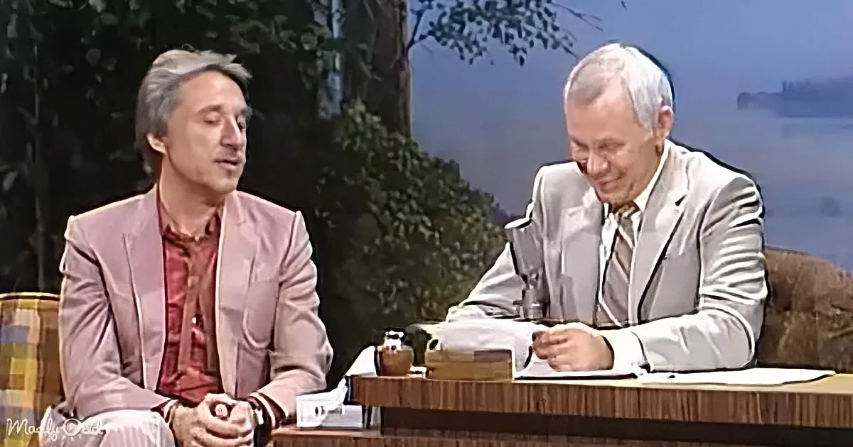Two men in suits are seated, one speaking while the other listens, with a scenic backdrop.