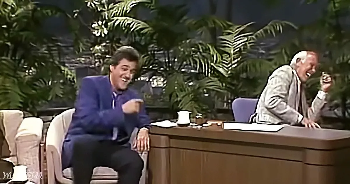 George Lopez and a guest are laughing on a talk show set with plants in the background.
