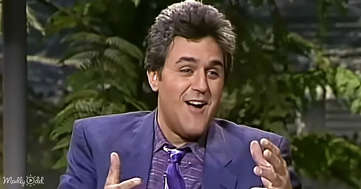 A man in a blue suit and striped shirt gestures with his hands, smiling, with green foliage in the background.