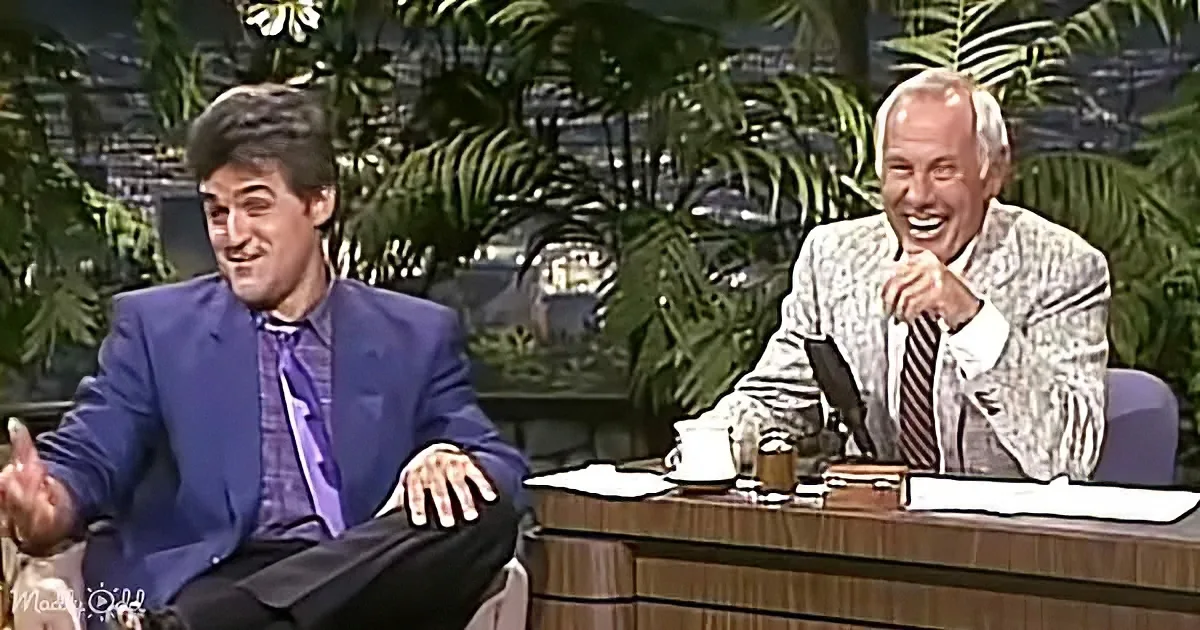 Two men, one in a blue suit, are seated on a talk show set with a backdrop of plants.