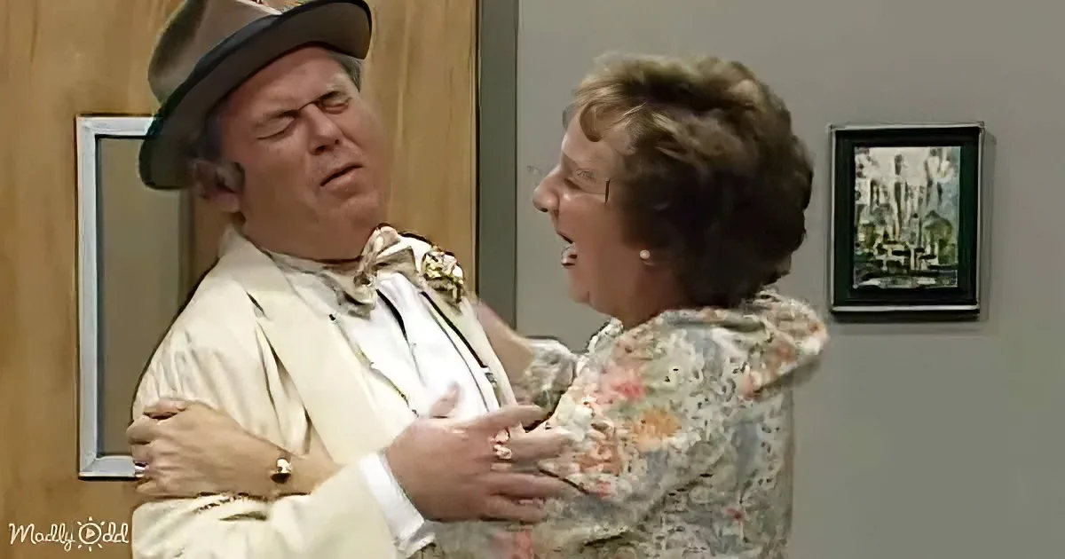 A man in a hat and white suit is embraced by a woman in a floral dress in a room with framed pictures.