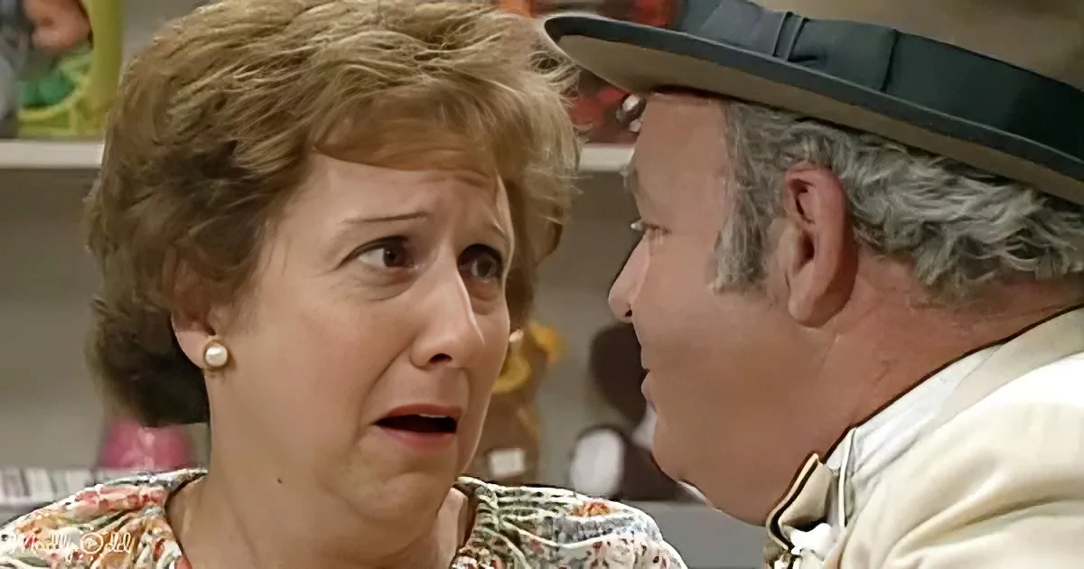 An older woman with short brown hair and pearl earrings looks surprised, while a man in a hat leans in close.