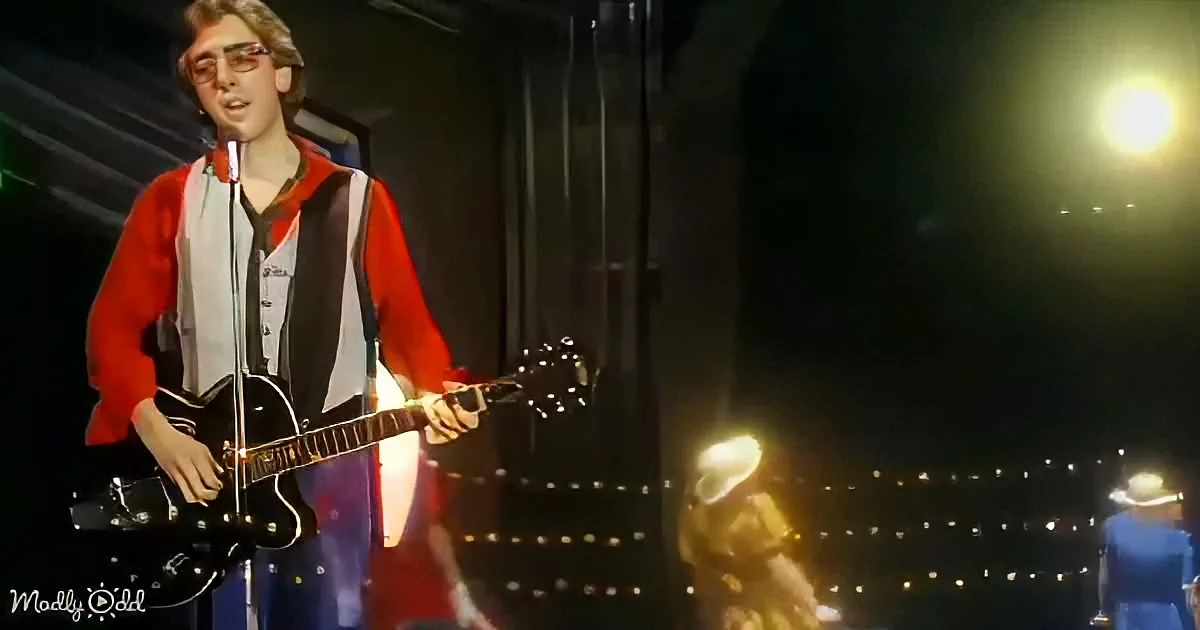 A person in a red and white outfit plays a black guitar on stage with a microphone and a bright light in the background.