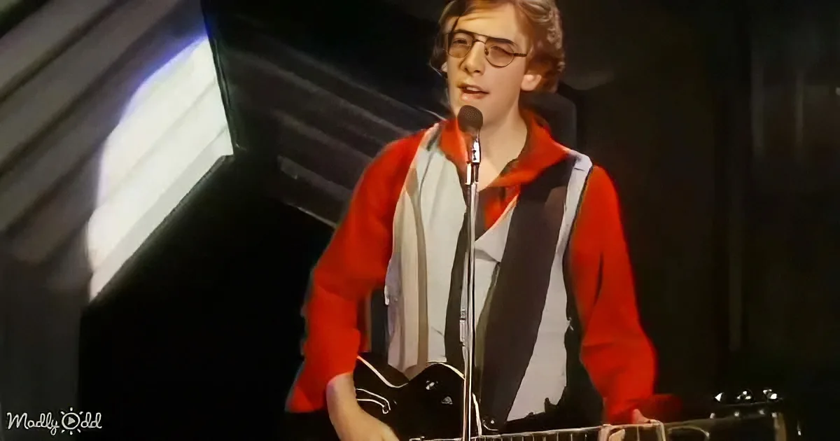 A person in a red and white outfit stands on stage, holding a guitar and singing into a microphone.