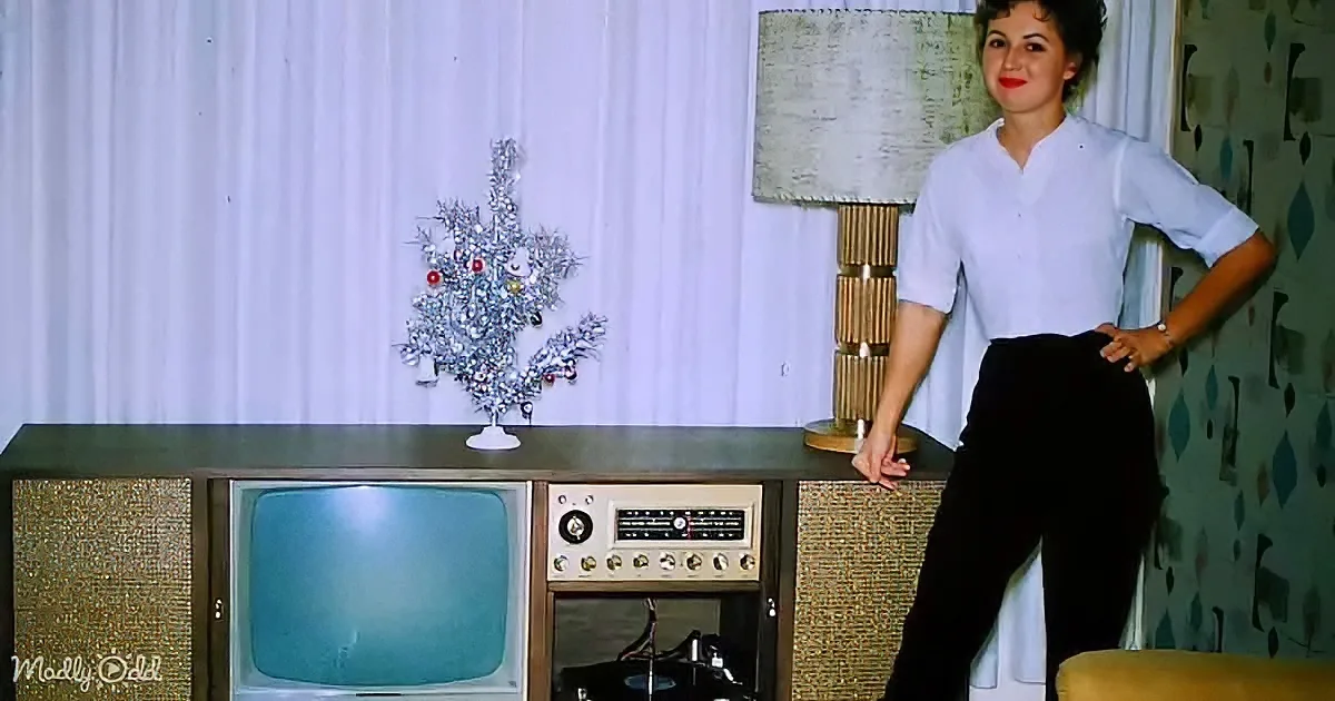 A woman in a white blouse and black pants poses beside a vintage TV and record player, with a small Christmas tree on a...