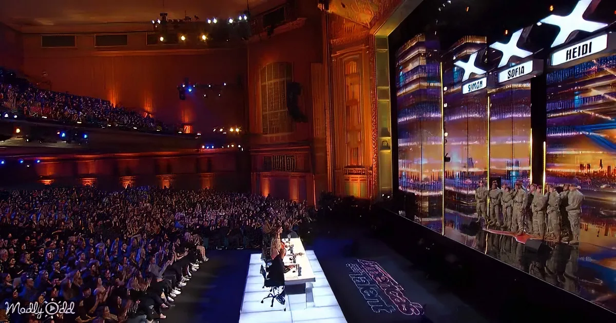 A fashion show with judges seated at a table, models on a runway, and a large audience in a grand theater setting.