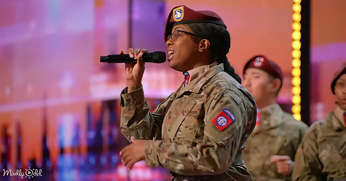 A person in military uniform sings into a microphone on stage, wearing a beret and glasses.