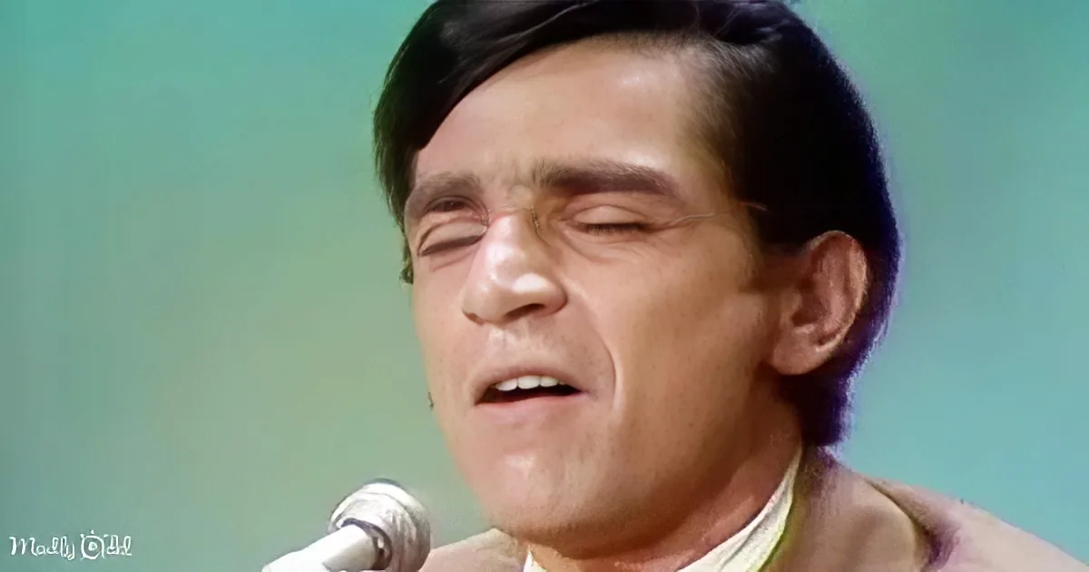 A man with dark hair and a light-colored shirt is singing into a microphone.