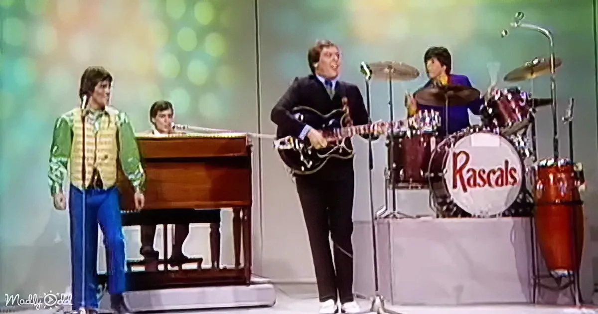 A man in a dark suit plays guitar on stage with a band, including a drummer and a pianist.