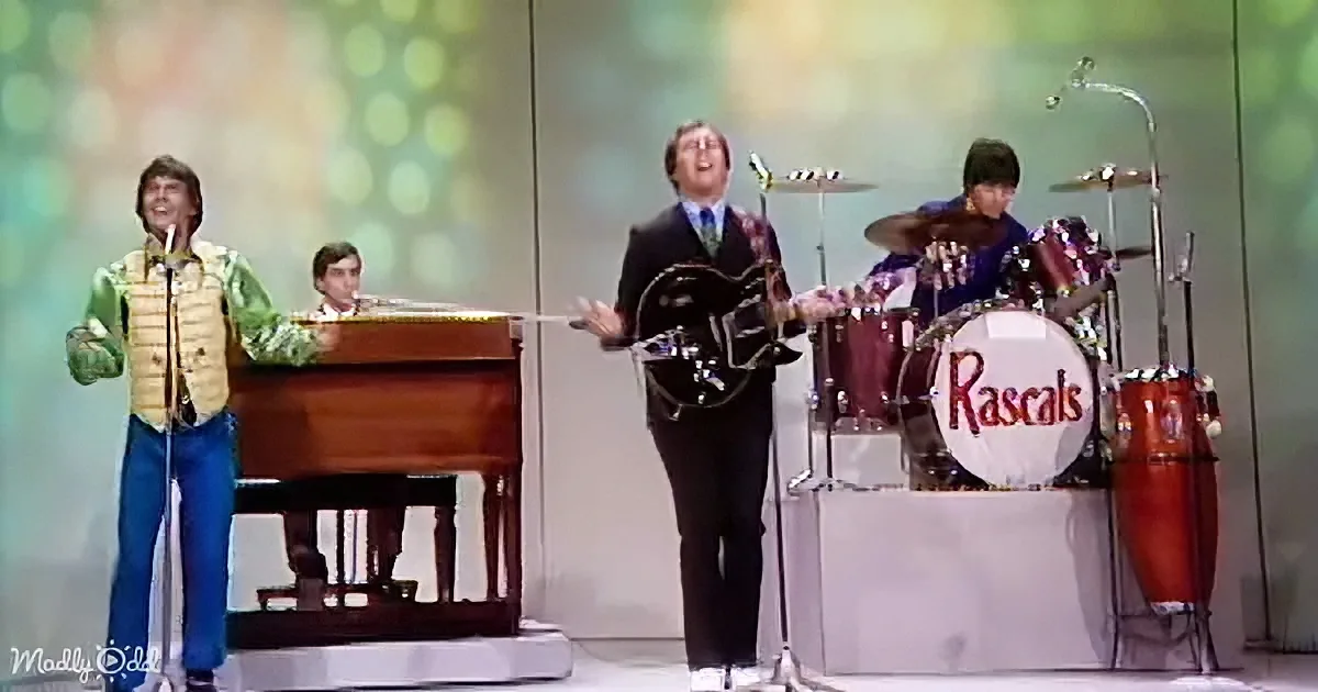 A man in a suit plays guitar on stage with a band, including a drummer and a piano player.