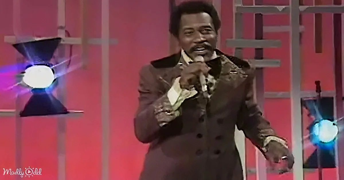 A man in a brown suit holds a microphone, gesturing with his other hand on stage.