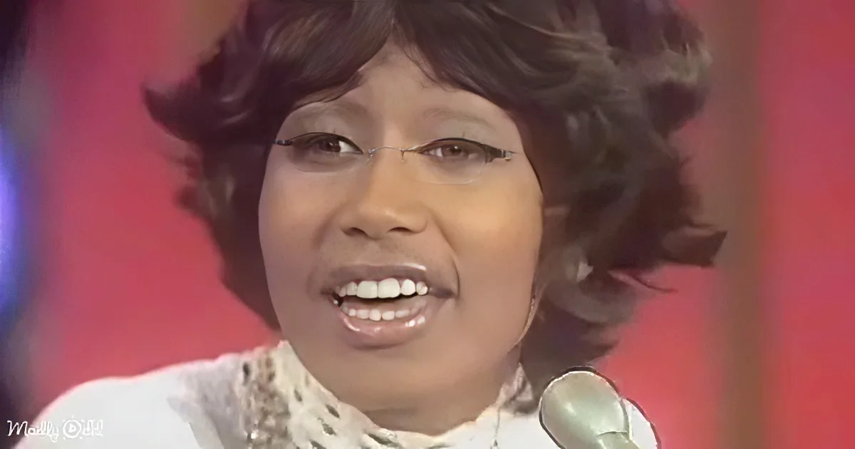 A smiling woman with short, curly hair and a white outfit is singing into a microphone.