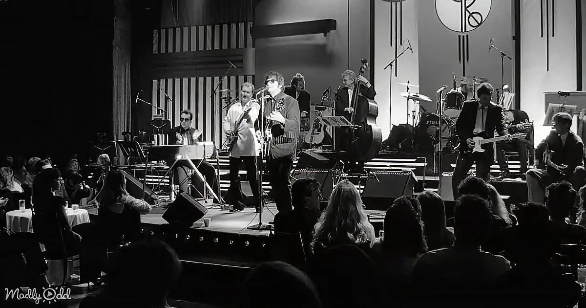 A band performs on stage with microphones and instruments, surrounded by an audience in a dimly lit venue.
