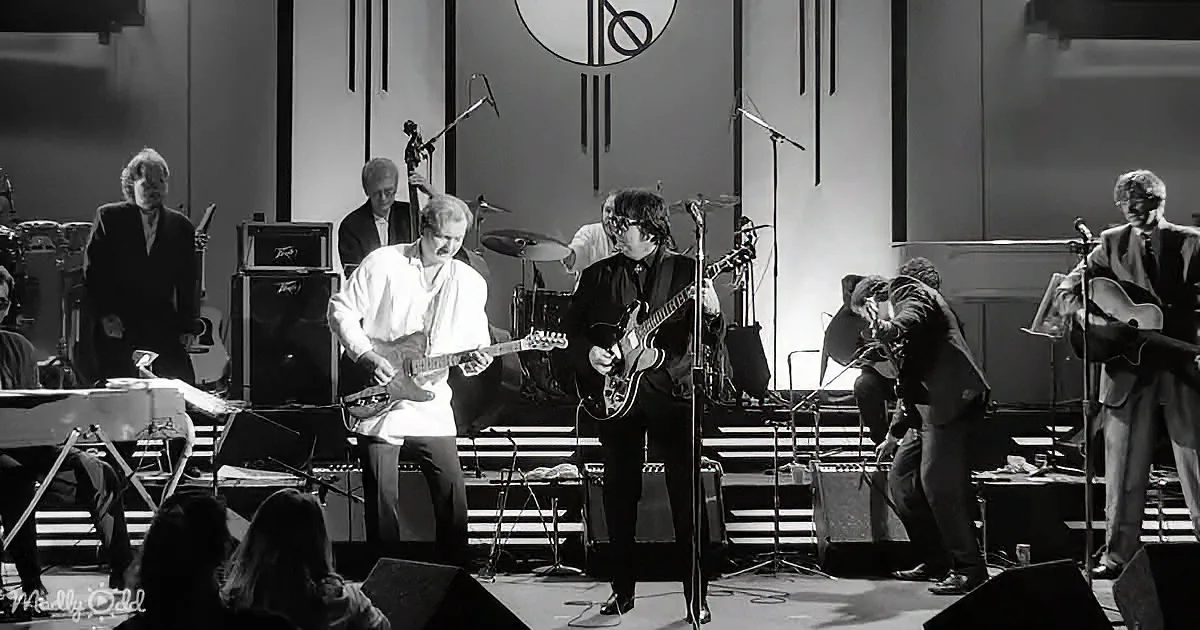 A guitarist in white shirt plays electric guitar on stage with other musicians and instruments.