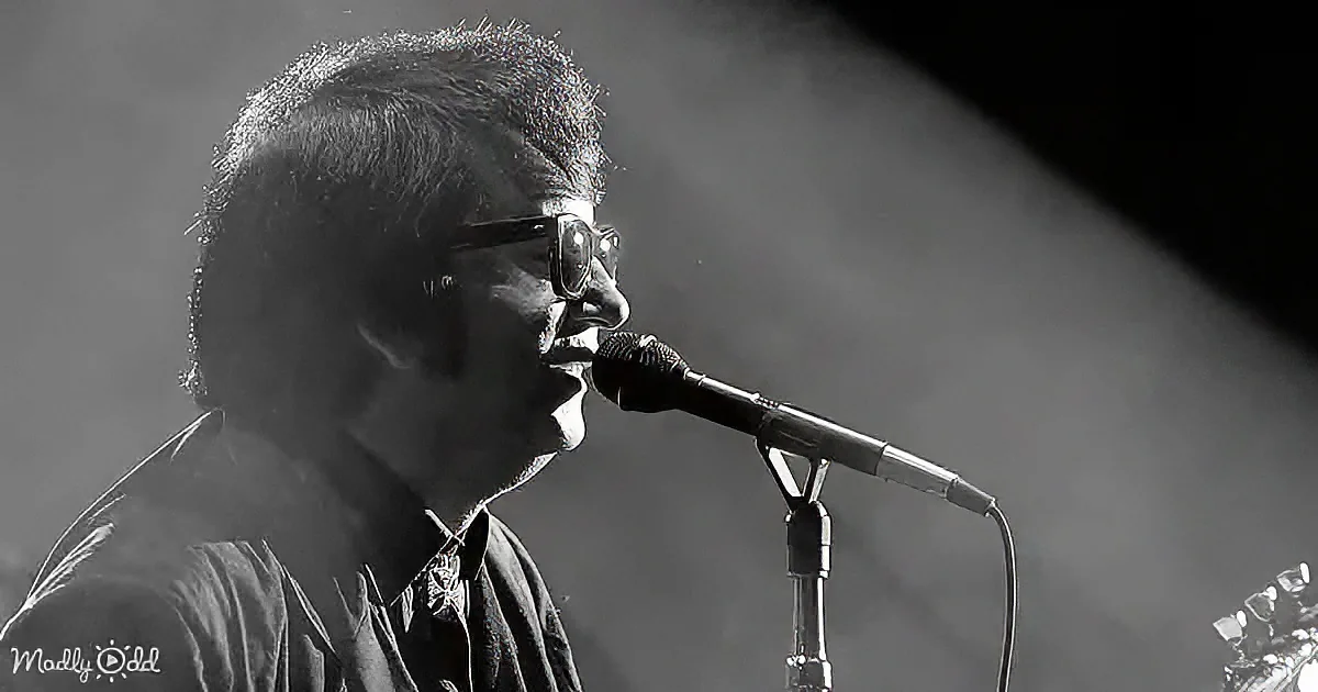 A man in glasses sings into a microphone on stage, illuminated by a spotlight.