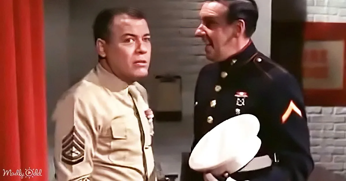 A man in a tan military uniform stands facing another in a dark uniform holding a white hat.