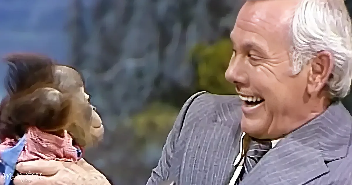 A man in a suit laughs while holding a puppet with a surprised expression.