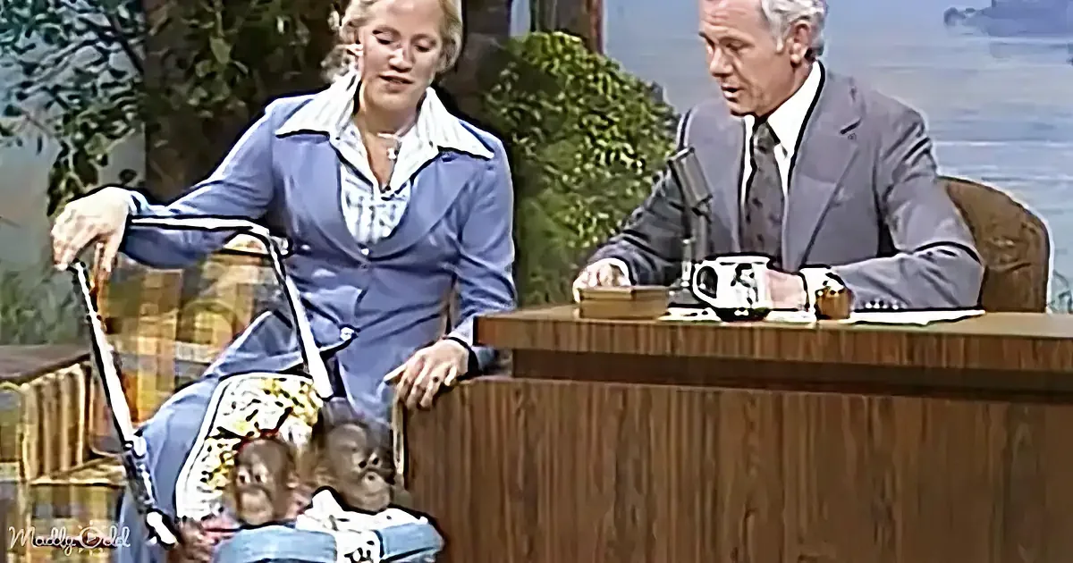 A woman in a blue suit leans on a desk, with a man in a suit sitting behind her, and two monkeys in a stroller in front...