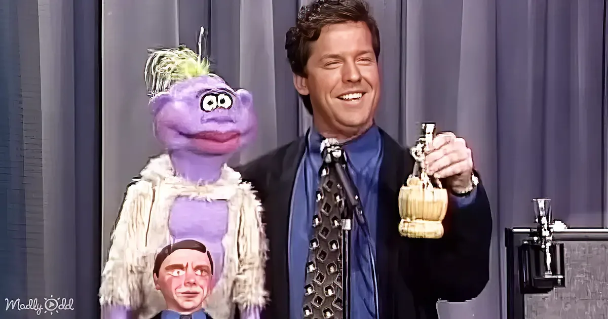 A man in a suit holds a trophy, smiling, with a purple puppet and another puppet below him.