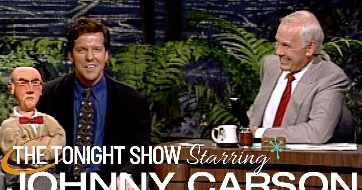 A man in a suit sits on a talk show set, smiling, with a puppet and another man in a suit behind him.